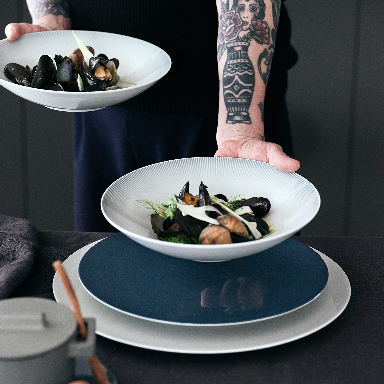 Une personne avec des tatouages sur le bras sert un plat de fruits de mer avec l'assiette à soupe en relief de TAC Sensual.